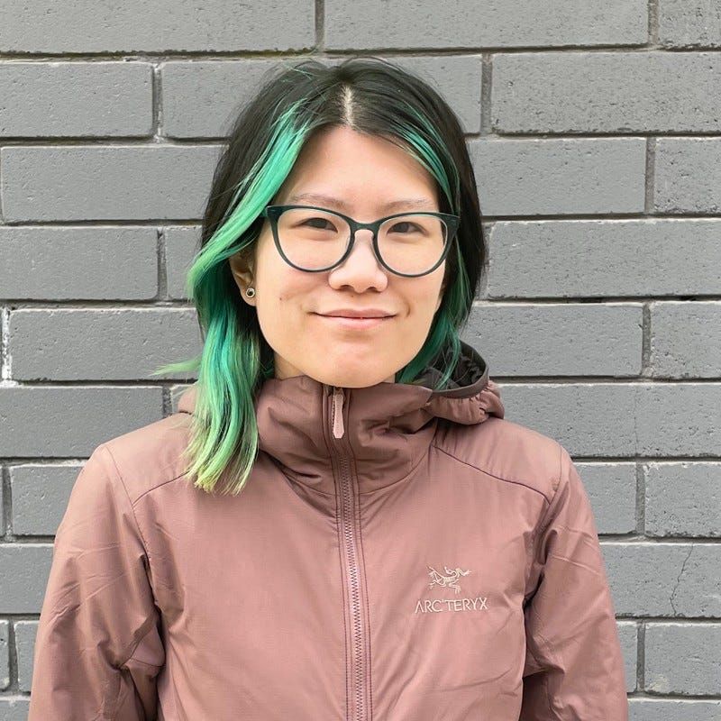 A picture of Jen Chan with a slight smile, she has dark brown hair with green accents and is wearing a coral winter coat. Behind her is a brick wall.