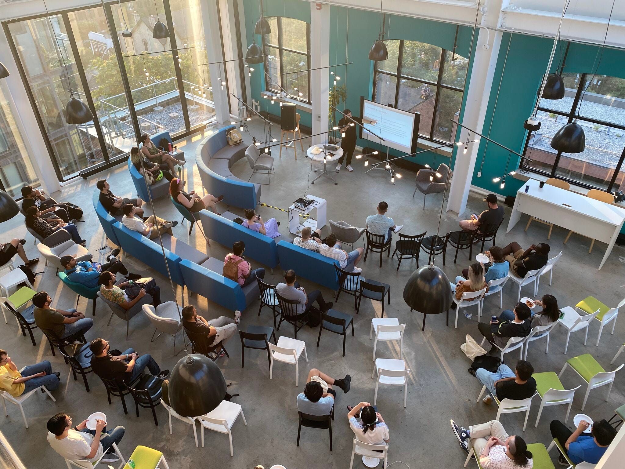 A top down photo of a room with people seated in chairs and couches forming a semi circle. In front of the people there is a speaker pointing to a presentation. The room is well lit with big windows on the background and lots of sun light coming in.