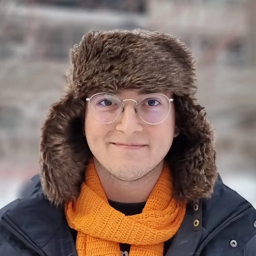 A picture of my face with a smile looking at the camera. I'm wearing aviator glasses, a fake fur winter hat and an orange scarf.